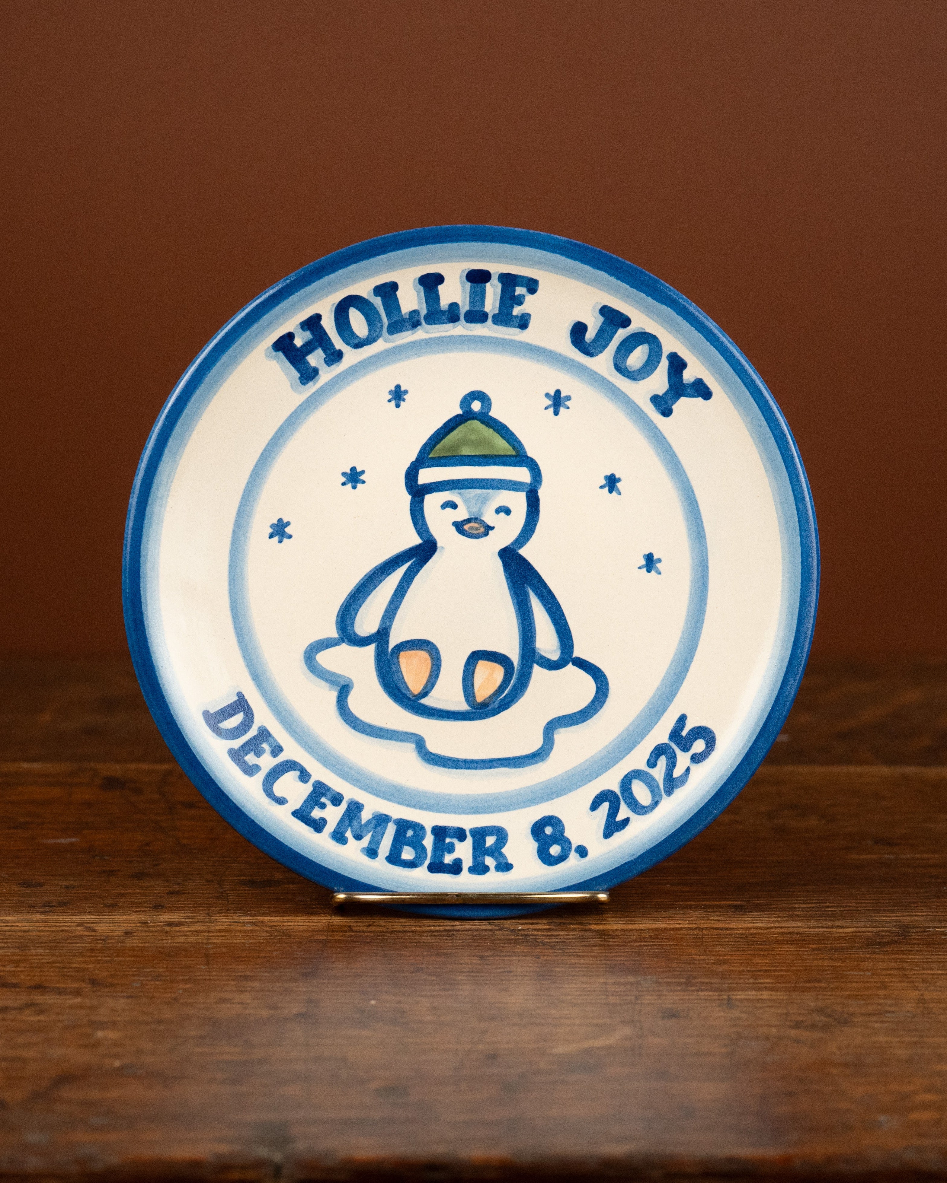 A round ceramic plate with a blue rim and a white center, featuring a blue penguin illustration and the text 'HOLLIE JOY' and 'DECEMBER 8, 2025', displayed on a wooden surface against a brown background.