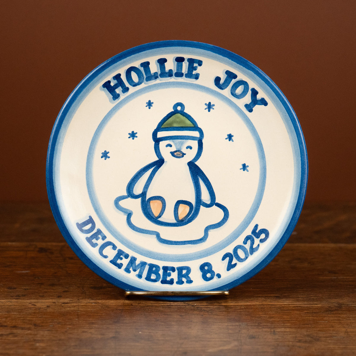A round ceramic plate with a blue rim and a white center, featuring a blue penguin illustration and the text 'HOLLIE JOY' and 'DECEMBER 8, 2025', displayed on a wooden surface against a brown background.