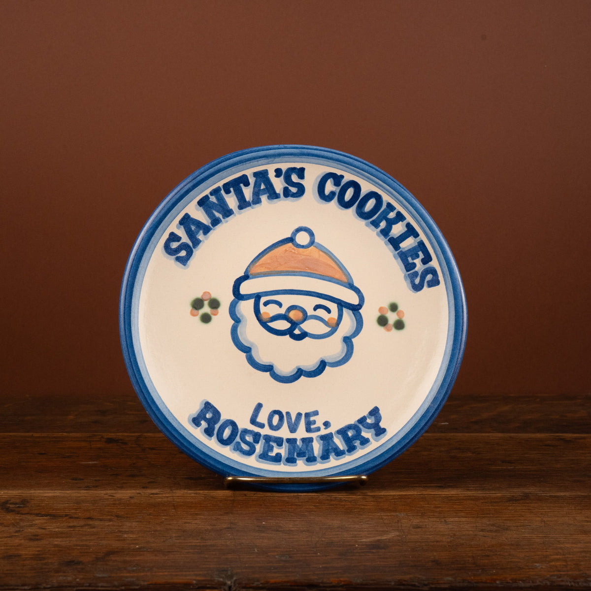 A round ceramic Christmas plate with a Santa cookie illustration and the text 'SANTA'S COOKIES' and 'LOVE: ROSEMARY', displayed on a wooden surface against a brown background.