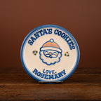 A round ceramic Christmas plate with a Santa cookie illustration and the text 'SANTA'S COOKIES' and 'LOVE: ROSEMARY', displayed on a wooden surface against a brown background.