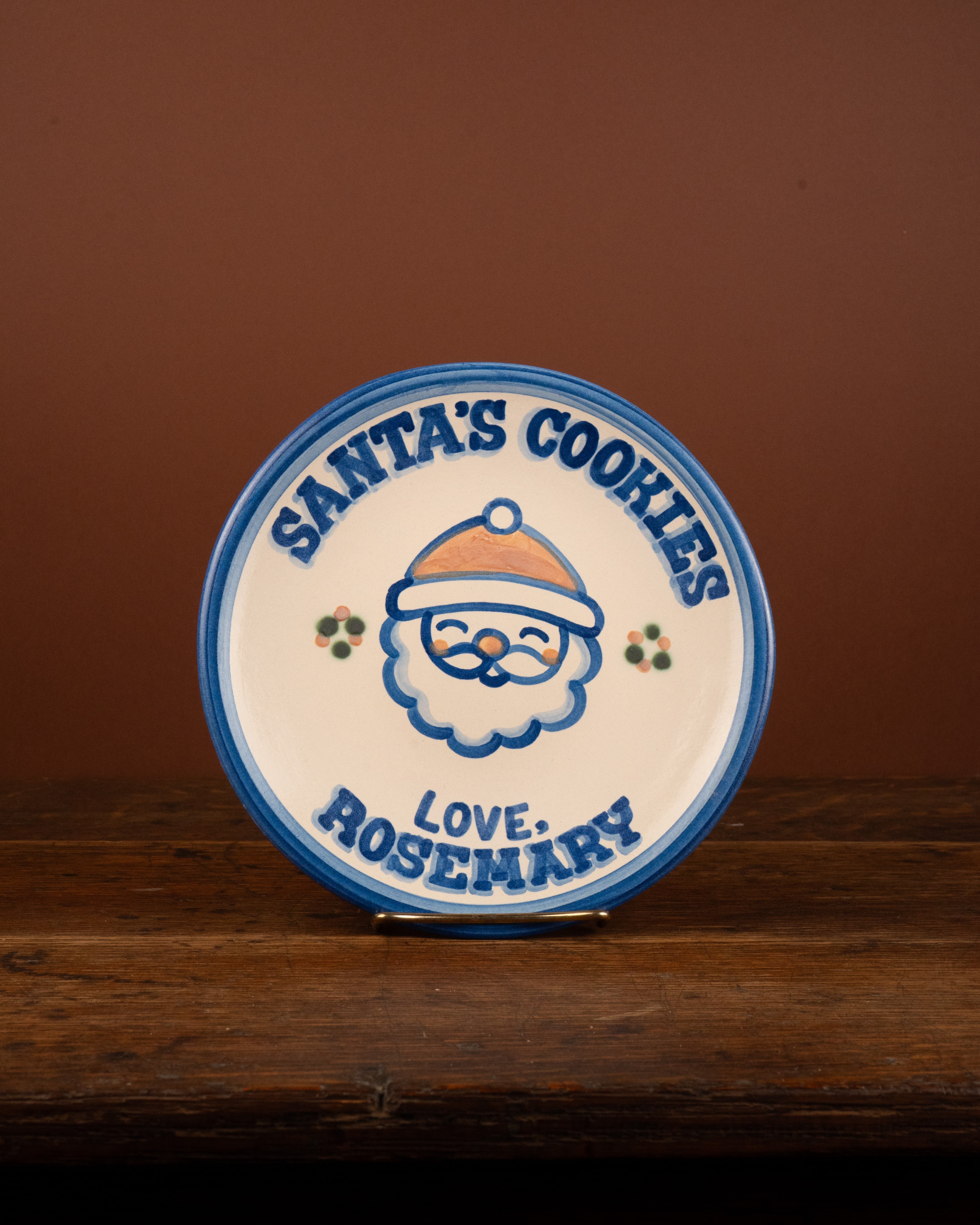 A round ceramic Christmas plate with a Santa cookie illustration and the text 'SANTA'S COOKIES' and 'LOVE: ROSEMARY', displayed on a wooden surface against a brown background.