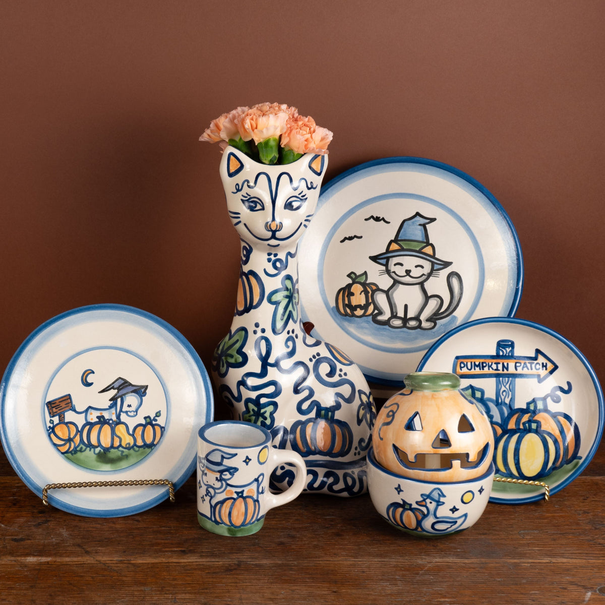 A set of ceramic Halloween-themed tableware including plates, a mug, a small bowl, and a tall vase, decorated with blue and orange illustrations of pumpkins, cats, and jack-o'-lanterns, arranged on a rustic wooden surface against a solid brown background.