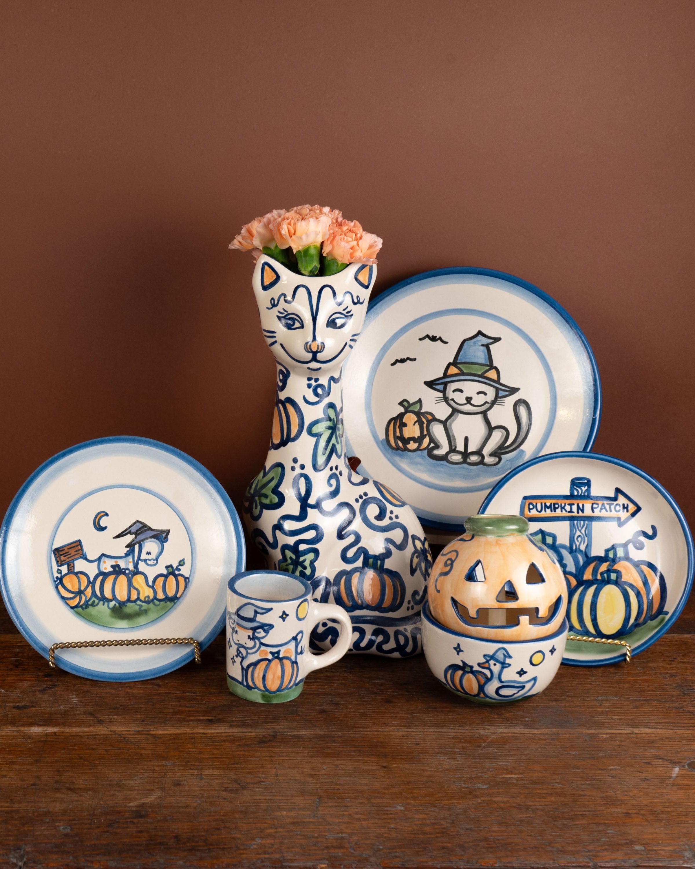 A set of ceramic Halloween-themed tableware including plates, a mug, a small bowl, and a tall vase, decorated with blue and orange illustrations of pumpkins, cats, and jack-o'-lanterns, arranged on a rustic wooden surface against a solid brown background.